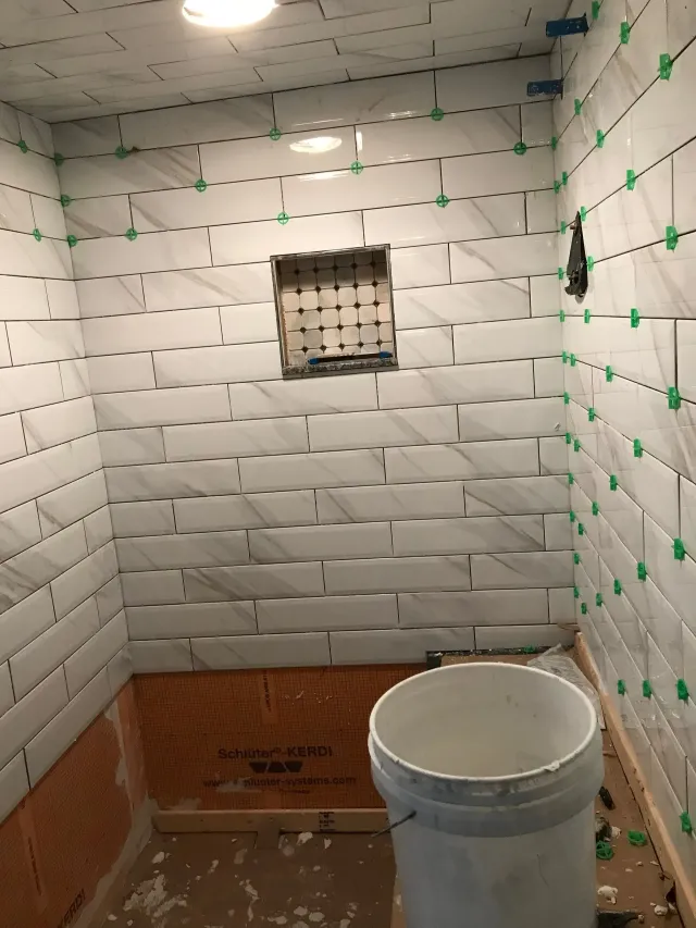 Small tiled shower stall under renovation, with white walls, exposed lower tile, and a bucket on the floor.